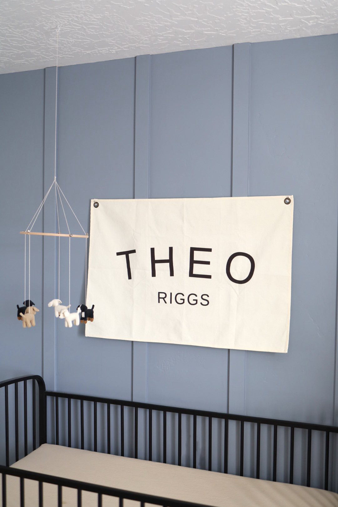 Custom canvas baby name banner with the name Theo Riggs hanging on a blue wall above a black crib