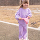 Young girl in a purple outfit riding a pink scooter on a paved path with a natural background.