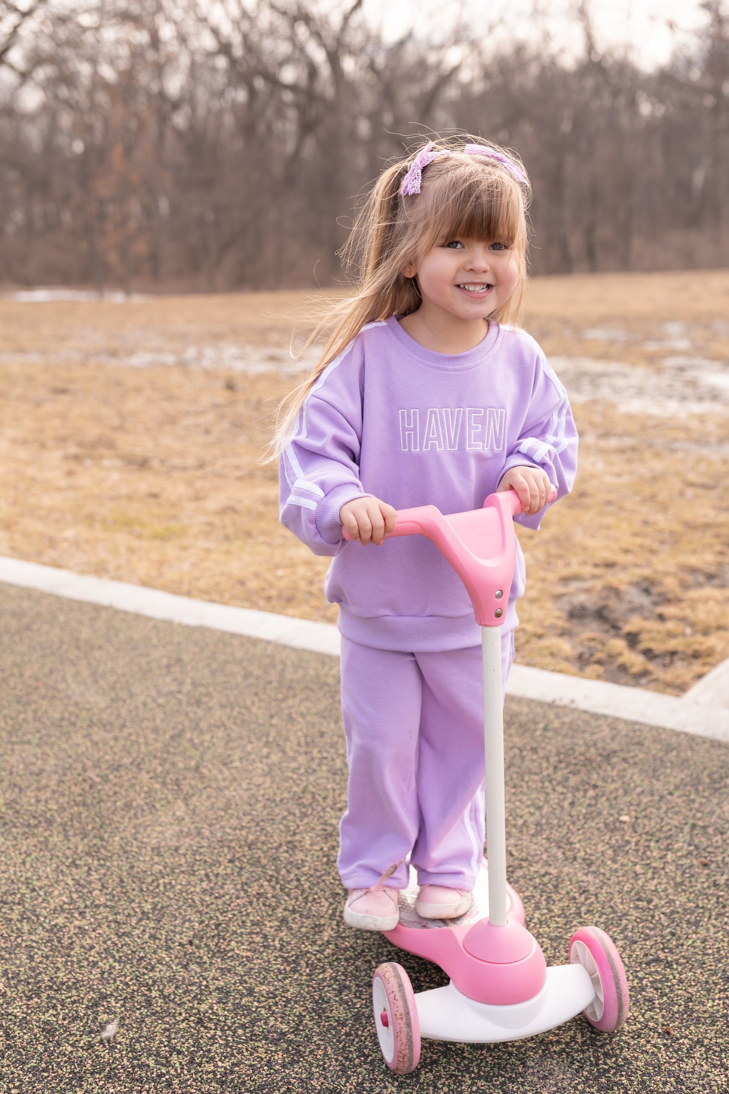 Young girl in a purple outfit riding a pink scooter on a paved path with a natural background.