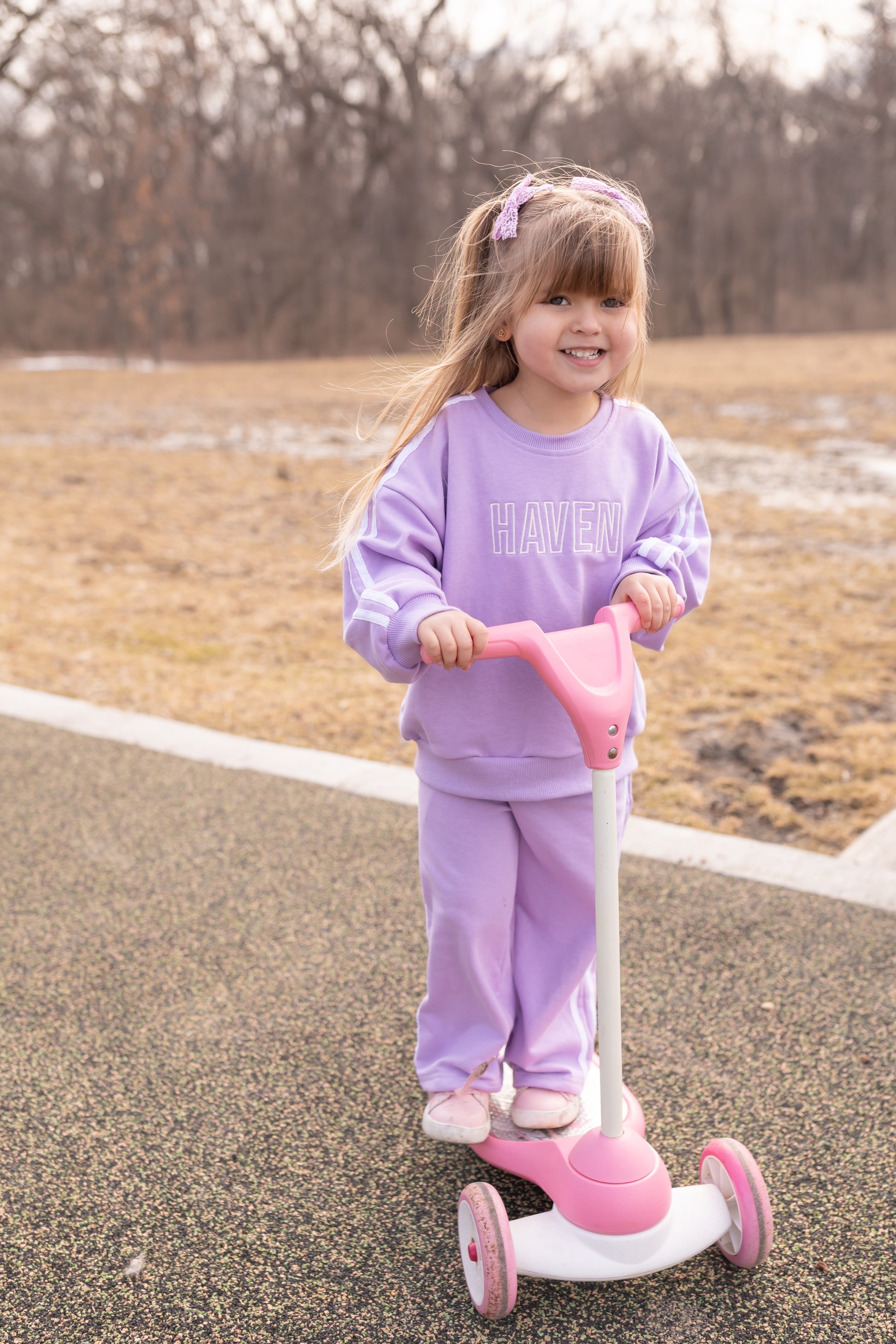 Young girl in a purple outfit riding a pink scooter on a paved path with a natural background.