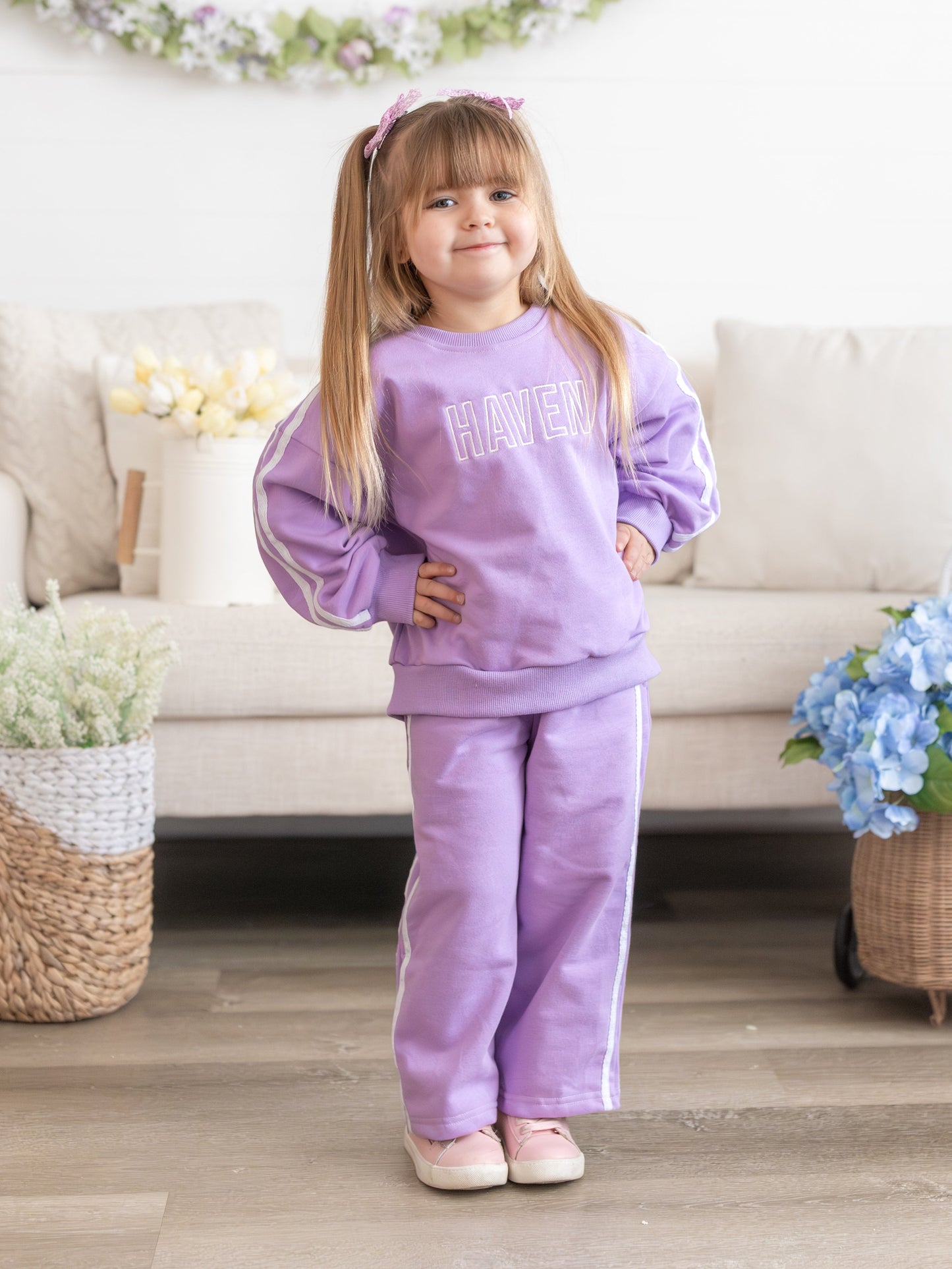 Young girl in a purple outfit standing in a living room with flowers and a couch.