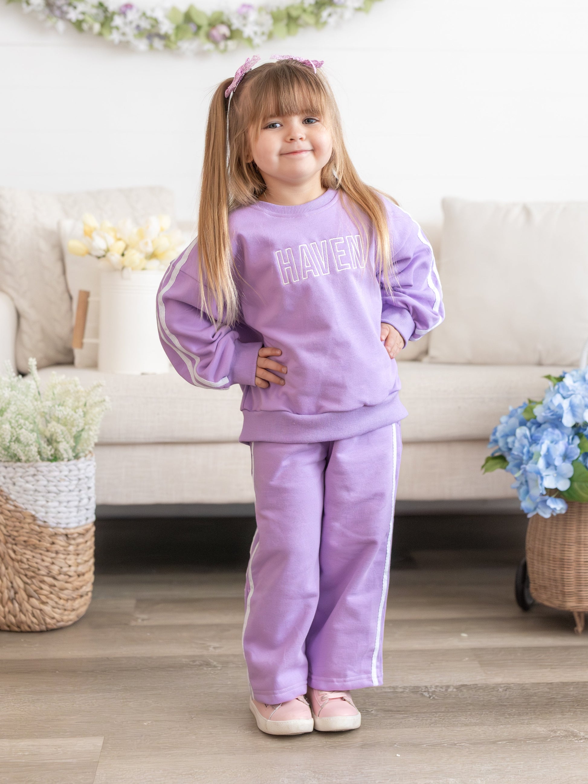Young girl in a purple outfit standing in a living room with flowers and a couch.