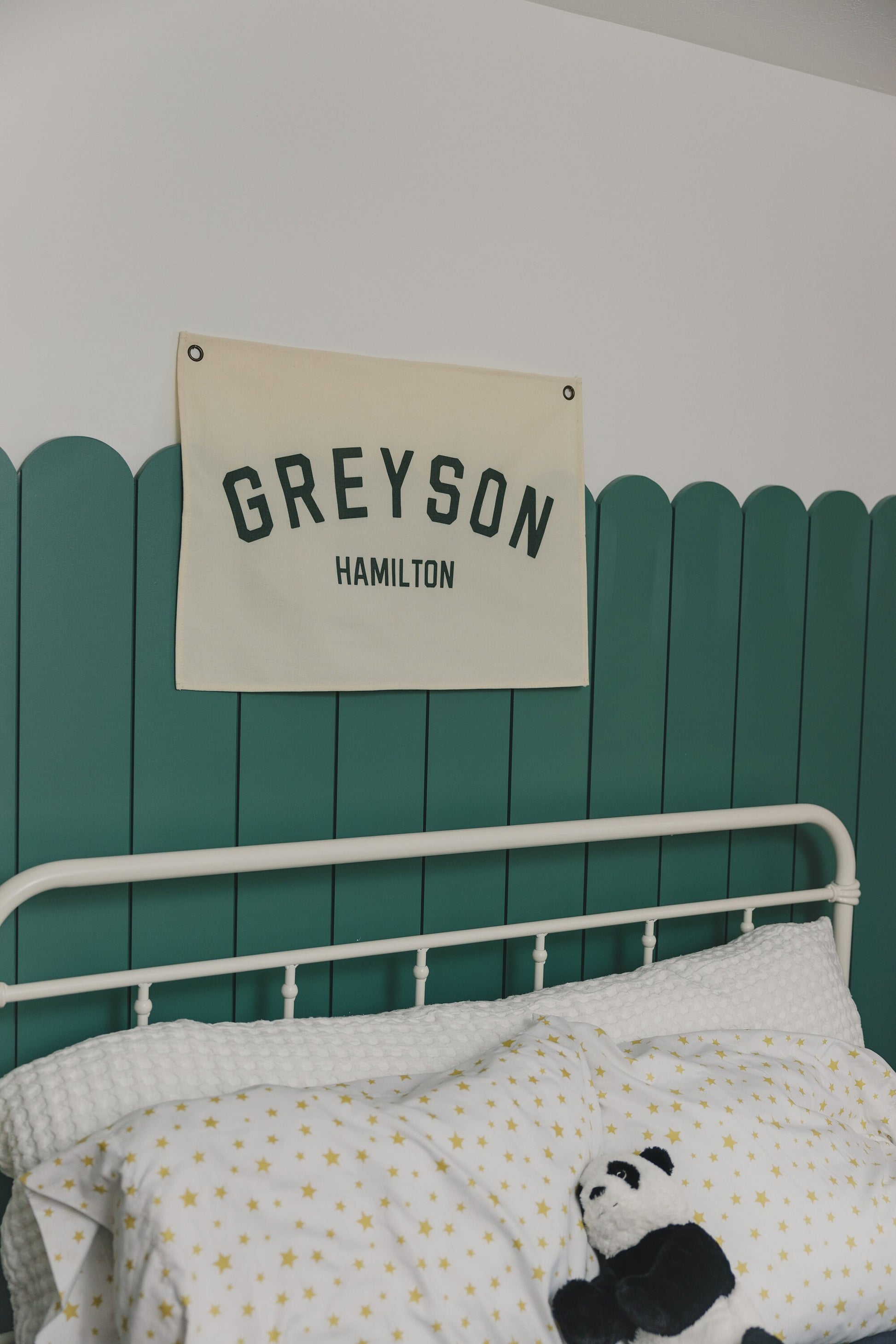 Custom canvas baby name banner with GREYSON HAMILTON hanging above white bed with star-patterned bedding and panda toy