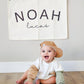 Custom canvas baby name banner with the name Noah Lucas hanging on wall behind smiling baby