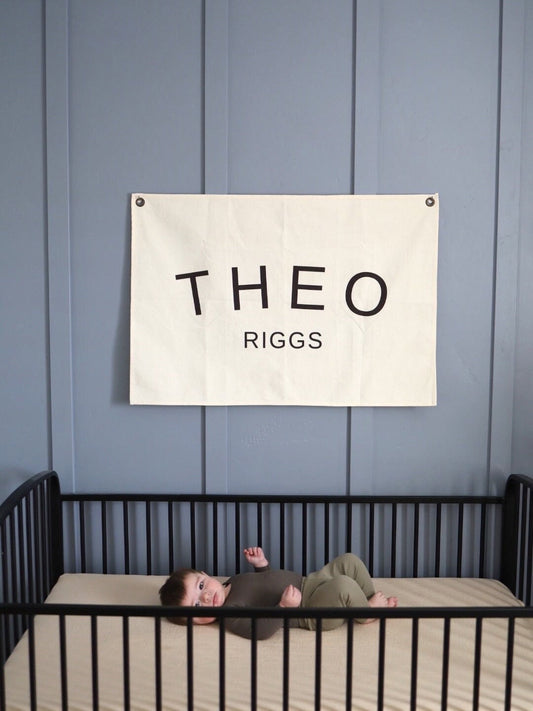 Custom canvas baby name banner with the name Theo Riggs hanging above a black crib